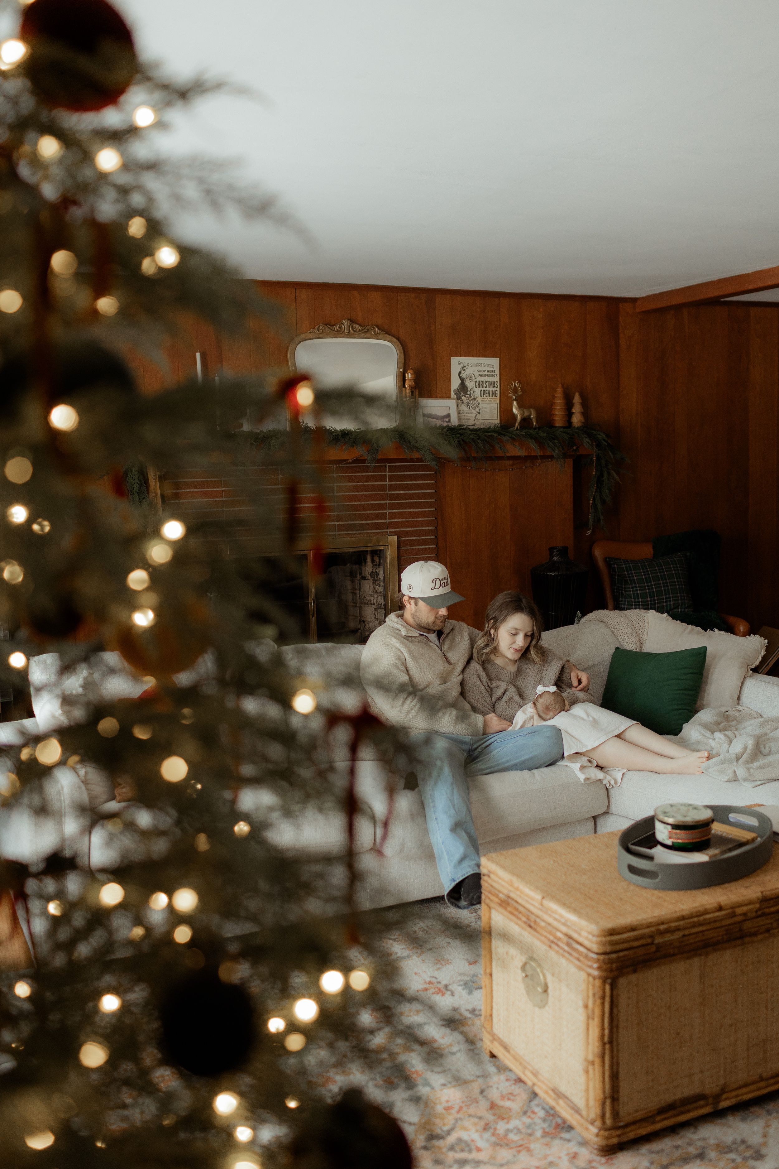 Couple sitting together on a cozy sofa beside a decorated Christmas tree with warm lights in a wood-paneled living room.
