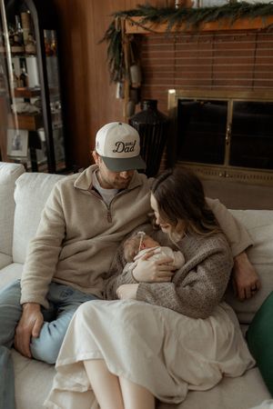 New parents sit on a cozy sofa, dad wearing a ‘Best Dad’ cap, lovingly gazing at their newborn baby.