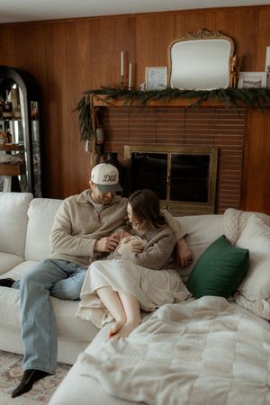 Couple sitting on white couch with newborn baby by cozy fireplace decorated with Christmas garland.