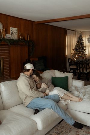 A couple sits on a white sofa cradling their newborn baby in a cozy living room decorated for Christmas.