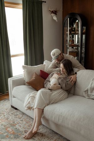 A couple sits on a white sofa, tenderly cradling their newborn baby in a cozy, warmly decorated living room.