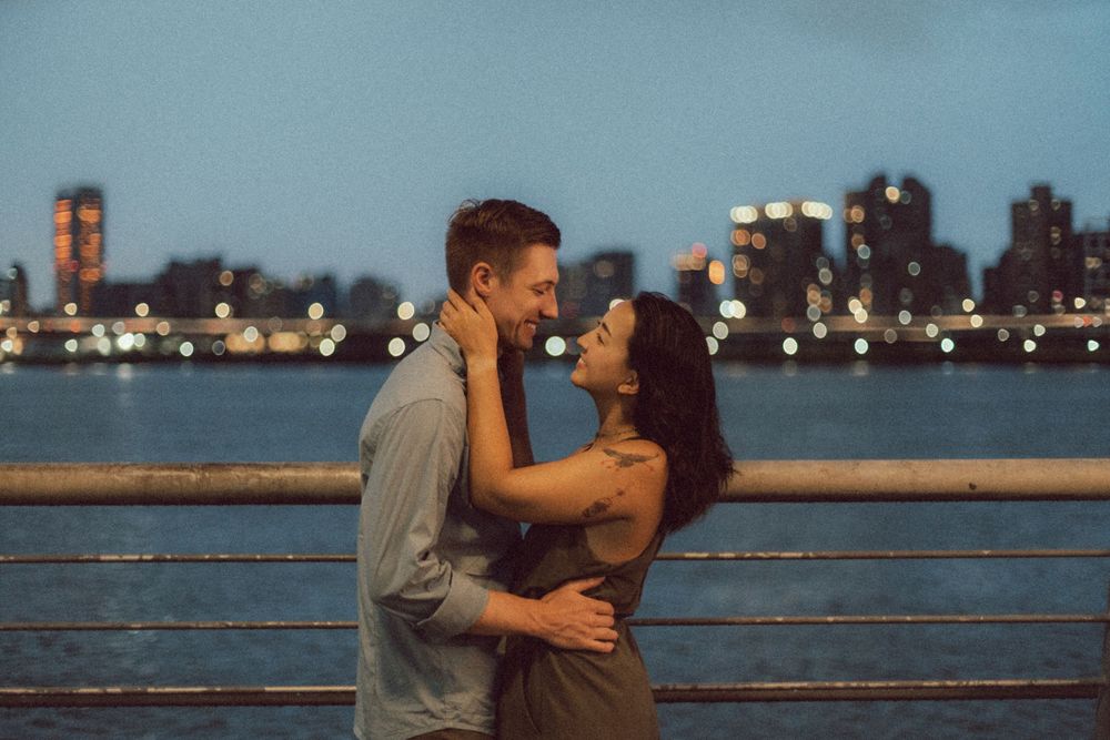 Romantic couple embracing at dusk on a waterfront pier with a glowing city skyline reflected on the river behind them.