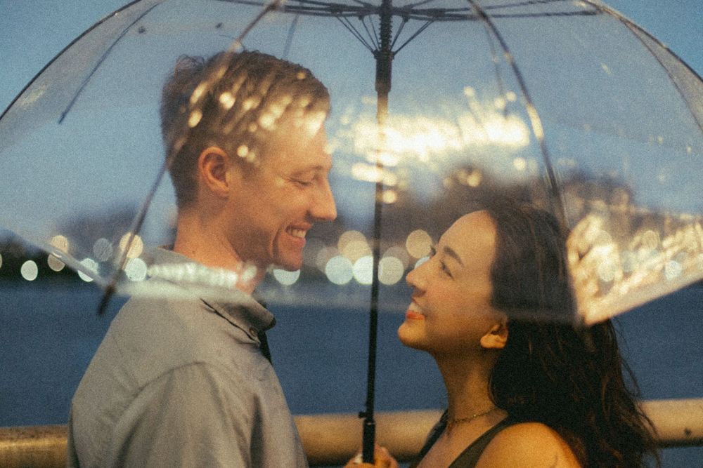 A smiling couple shares a romantic moment under a clear umbrella at dusk near a waterfront.