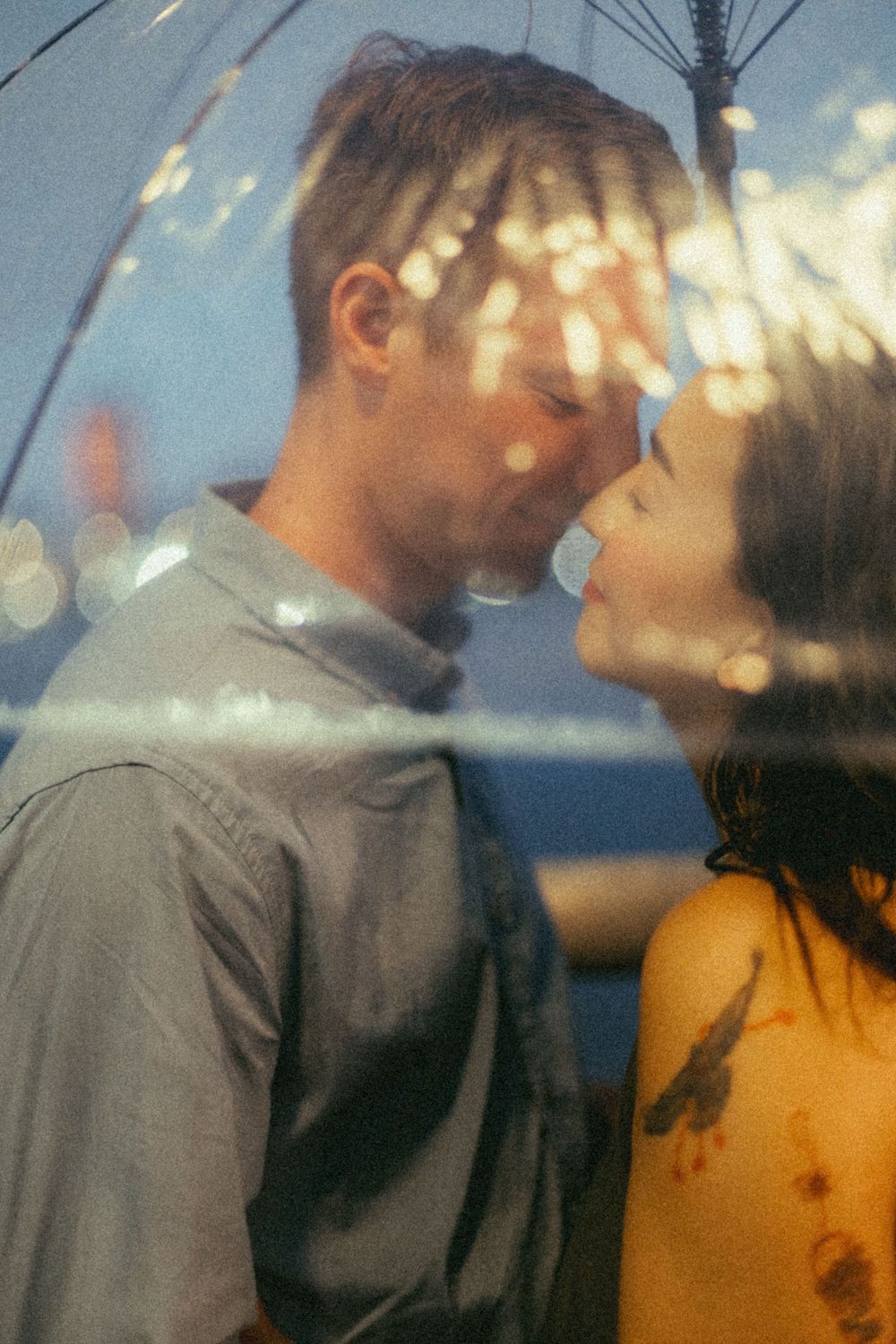 A romantic couple sharing an intimate moment under a clear umbrella with bokeh lights in the background.
