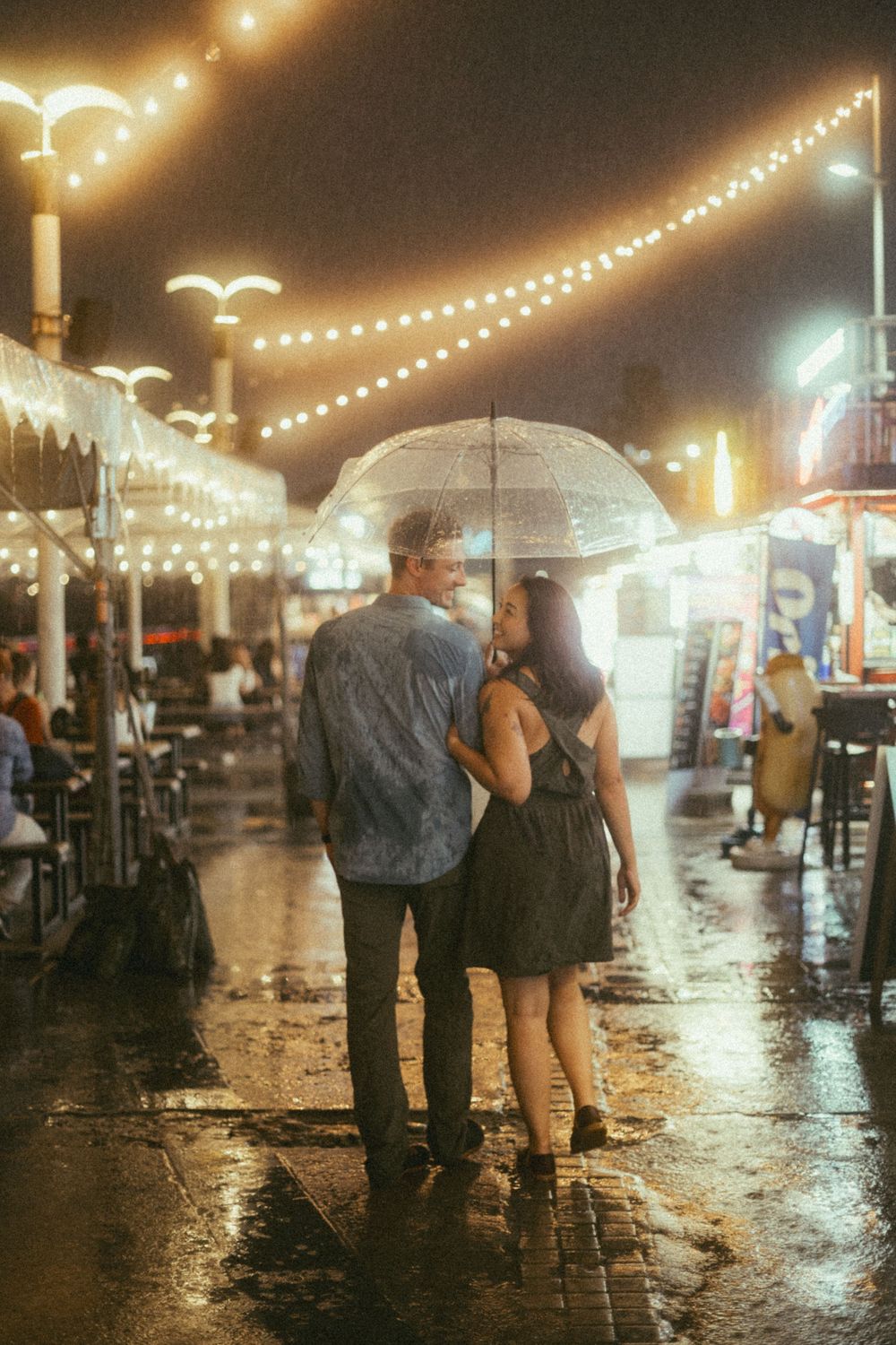 Couple sharing a clear umbrella walking together on a rainy night street with string lights glowing above.