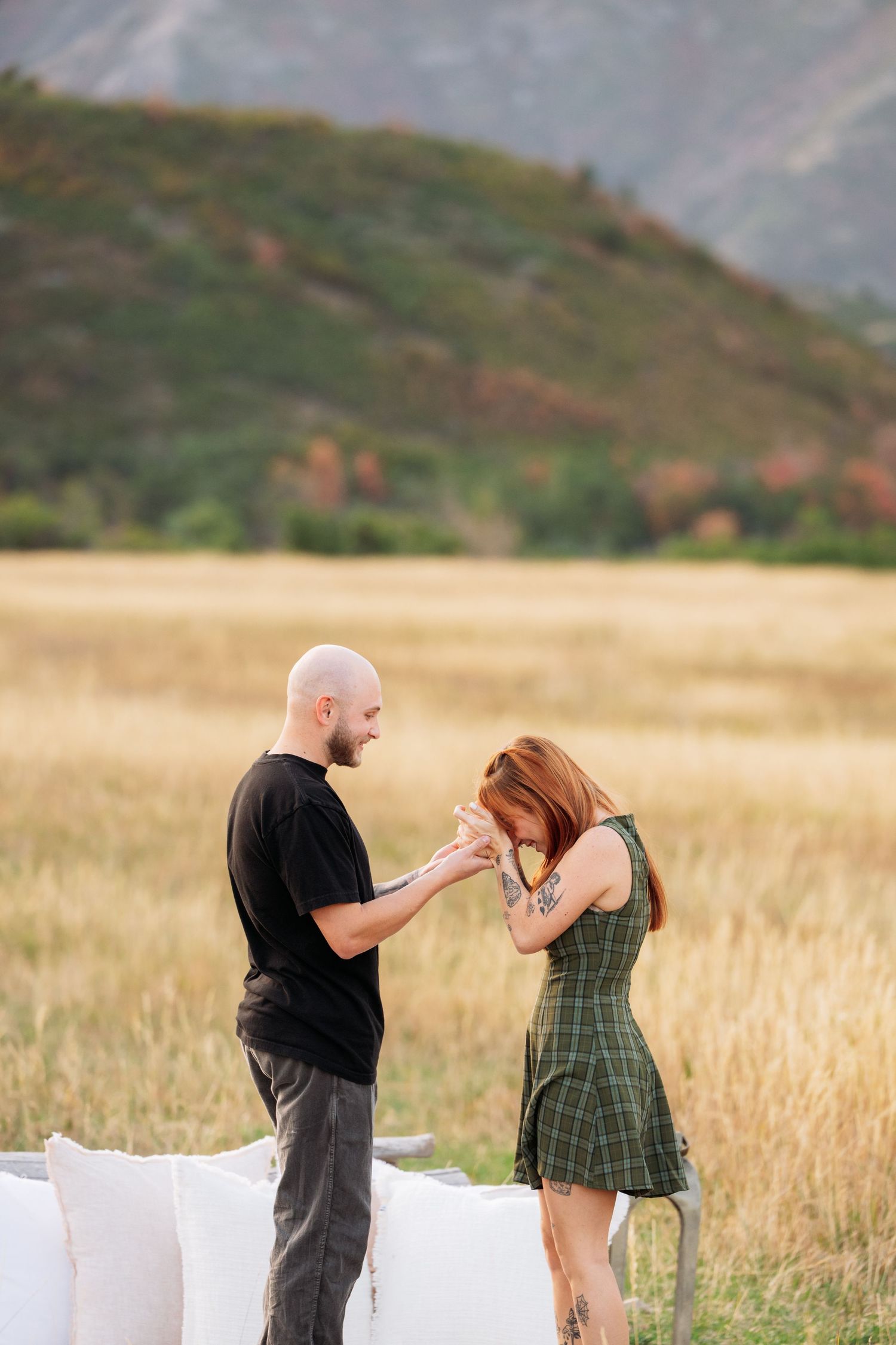A bald man in a black shirt holds a redhead woman's hands in a plaid dress in a golden field with mountains behind.
