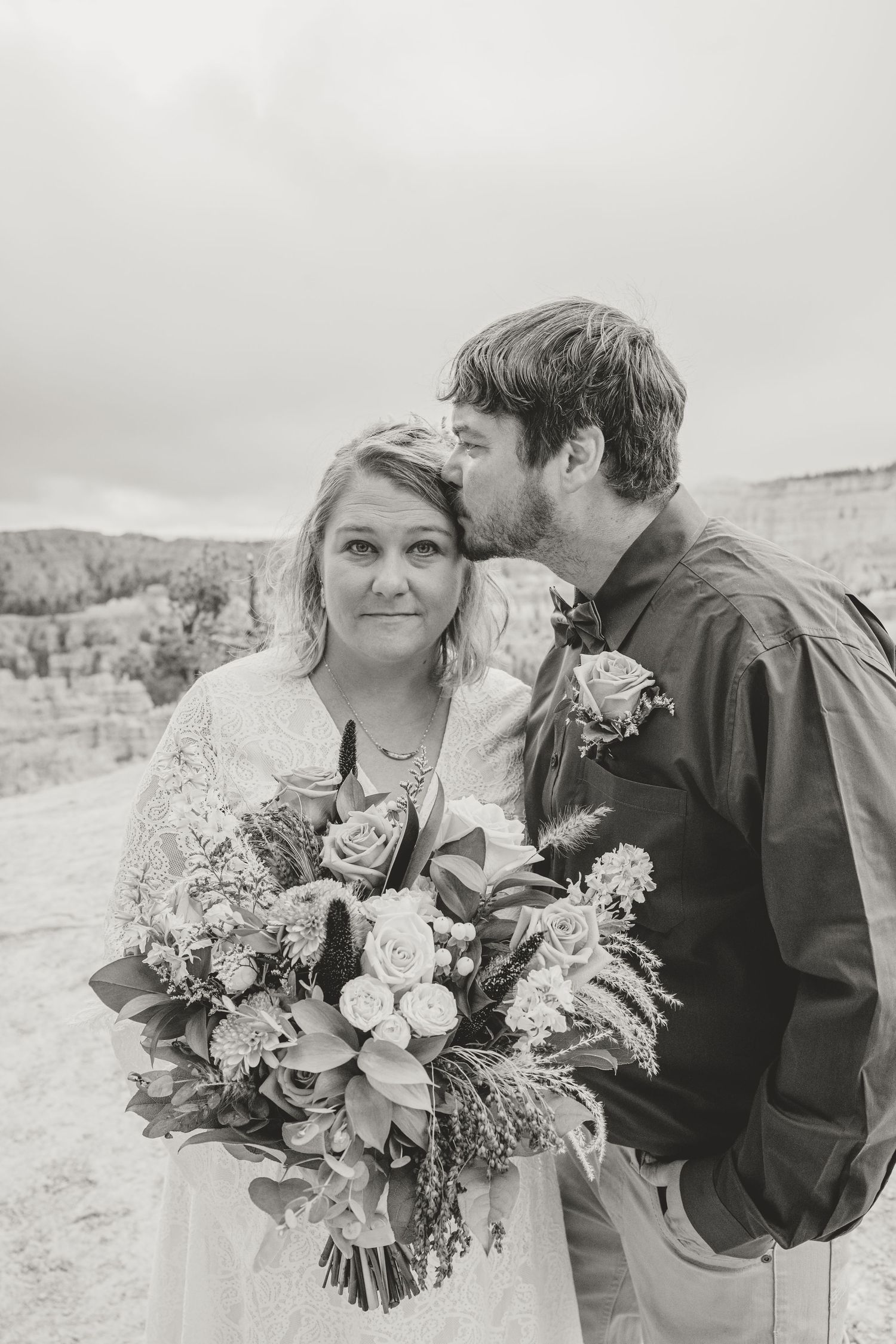 Black and white wedding photo of bride holding bouquet while groom kisses her cheek outdoors.