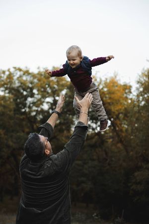 Smiling father tossing his laughing toddler into the air outdoors beneath autumn trees during a family photo session.