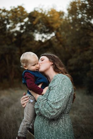Mother in a green floral dress kisses her toddler son on the cheek while holding him in a golden autumn field.