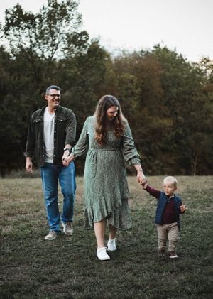 Parents hold hands and walk through a grassy field with their toddler son, surrounded by fall trees at dusk.