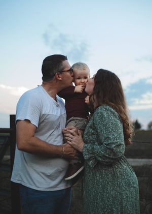 Mom and dad kiss their toddler son on both cheeks while standing outdoors under a soft pastel evening sky.