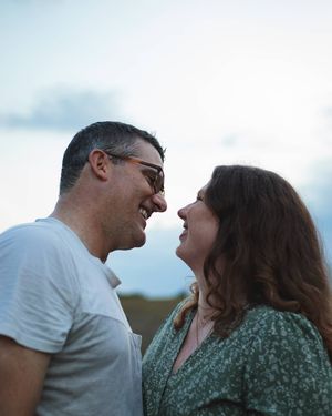 Smiling couple stands face to face with foreheads close in a romantic outdoor portrait against a cloudy blue sky.