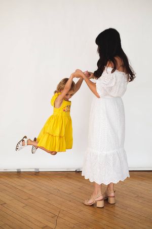 Mother in a white eyelet dress swings her giggling daughter in a yellow dress in a bright studio with wood floors.