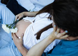 Expectant mother in a white top rests on a blanket holding white carnations on her baby bump as her partner leans in.