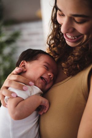 Smiling woman with curly hair cradles a laughing newborn baby against her shoulder in a warm, tender moment.
