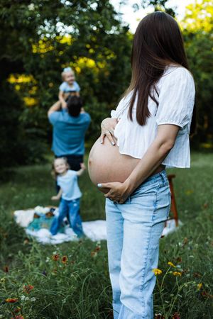 Pregnant woman in jeans holds her belly in a grassy park while a man lifts a toddler and another child stands nearby.