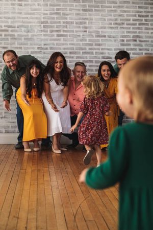 Family in colorful outfits crouches smiling on a wood floor as a toddler in a floral dress runs toward them.