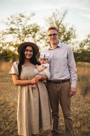 Smiling couple in a golden field holds their baby girl with a white bow, posing for a sunset family portrait.