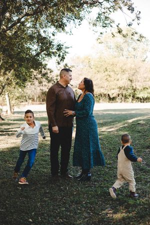 Parents embrace under a large tree while two young boys play on the grass during an outdoor autumn family photo session.