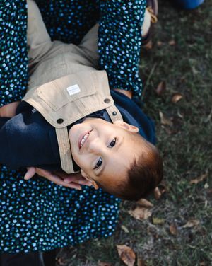 Smiling little boy in tan overalls looks up at the camera while being held by his mother on a grassy, leaf-covered ground.