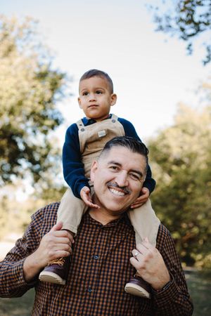 A smiling man with a mustache carries a toddler in tan overalls on his shoulders in a sunny park setting.