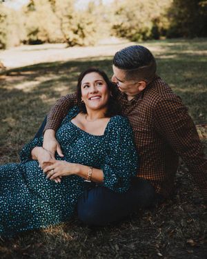 A smiling woman in a green floral dress sits on the grass while a man in a brown plaid shirt embraces her from behind.