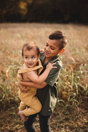 A smiling older boy in a green shirt hugs a laughing toddler in tan overalls in a golden grassy field.