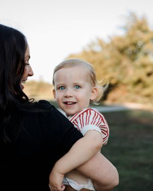 A smiling woman with long dark hair holds a happy blond toddler in a red striped shirt outdoors with trees behind them.