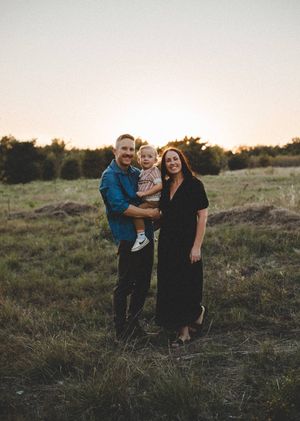 A smiling family of three, a man, woman and toddler, pose together in a grassy field at sunset.