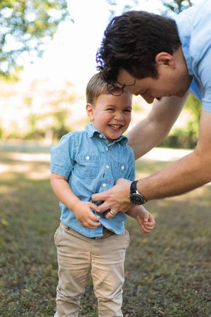 A laughing toddler in a blue chambray shirt and khaki pants plays outdoors as a dark-haired man leans down to tickle him.