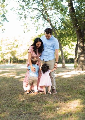 A smiling family of four embraces on a sunlit grassy lawn beneath tall trees during an outdoor family portrait session.