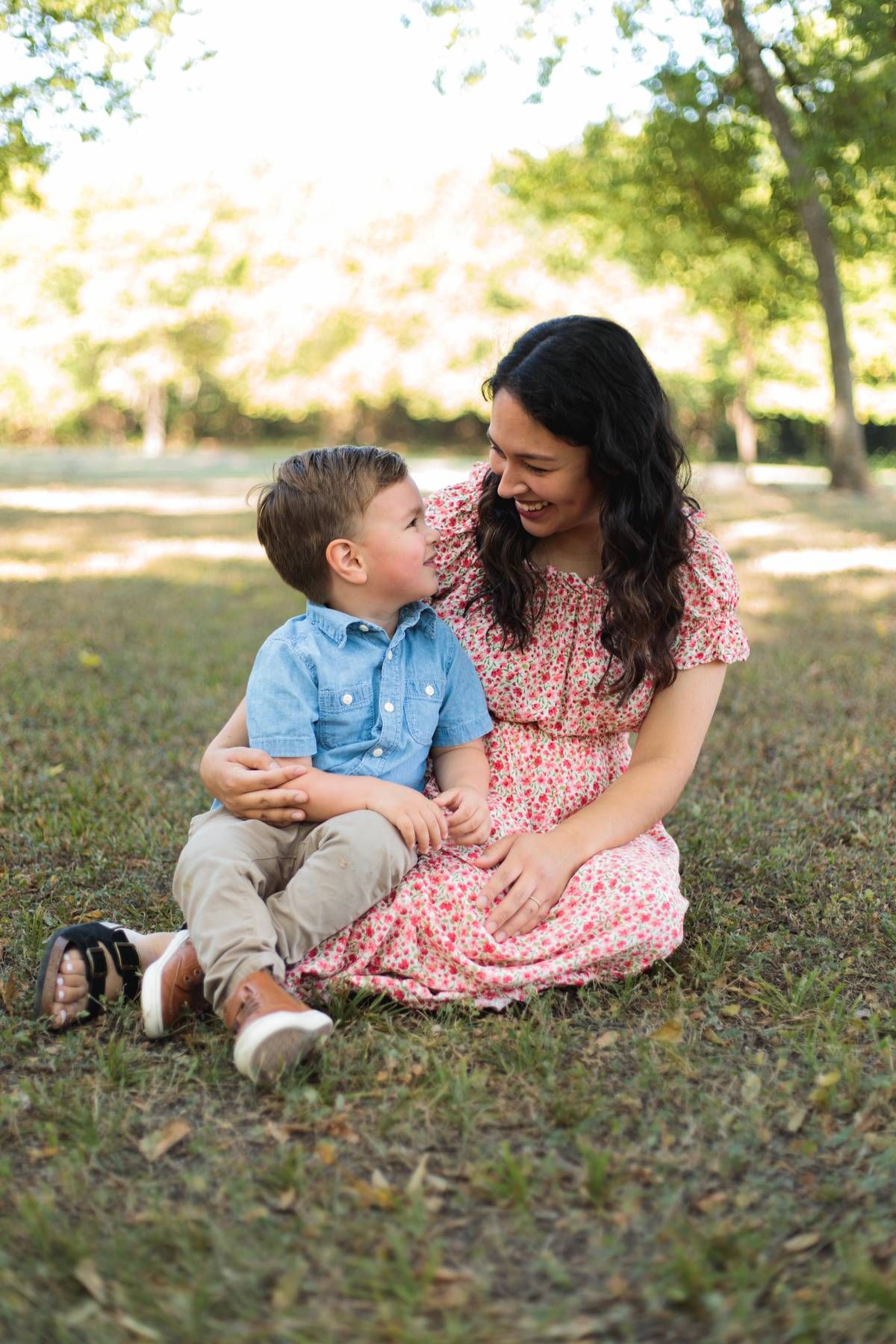 A smiling mother in a floral dress sits on the grass with her young son in a blue button-up shirt in a sunny park.