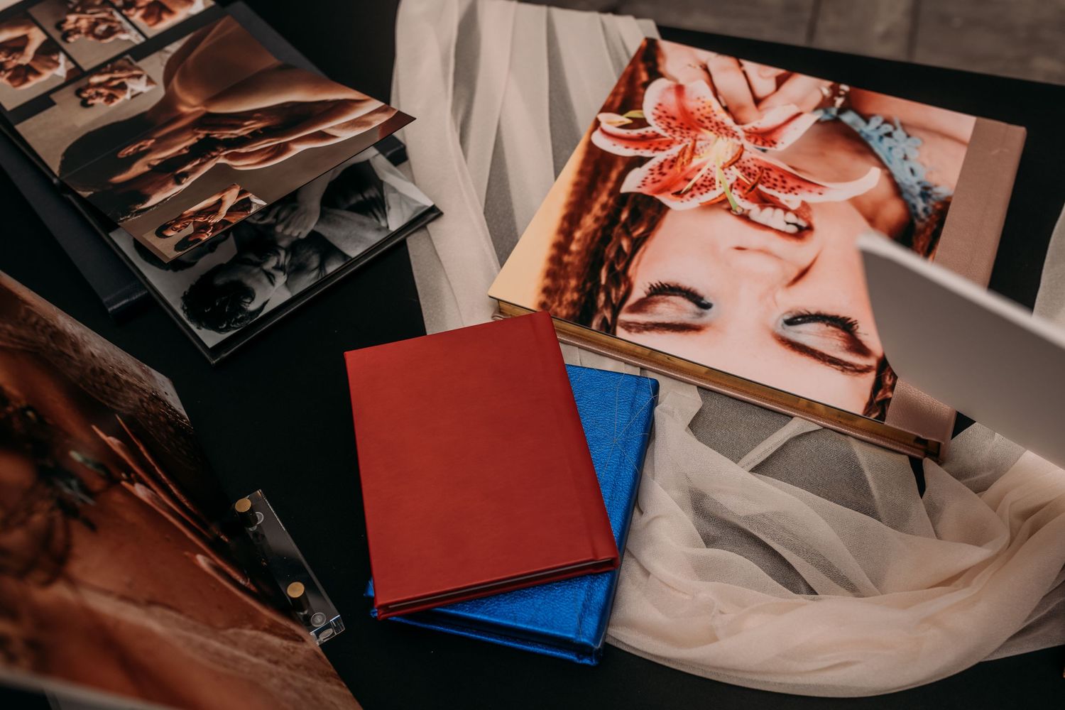 Red and blue photo albums beside printed portrait photographs and a proof book displayed on a dark table with white tulle fabric.