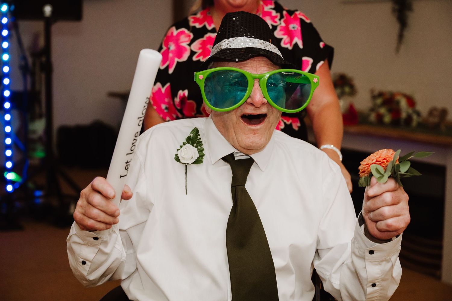 Grandad black hat, oversized green sunglasses and white boutonniere laughs while holding a light baton  at a festive wedding at Cott Farm