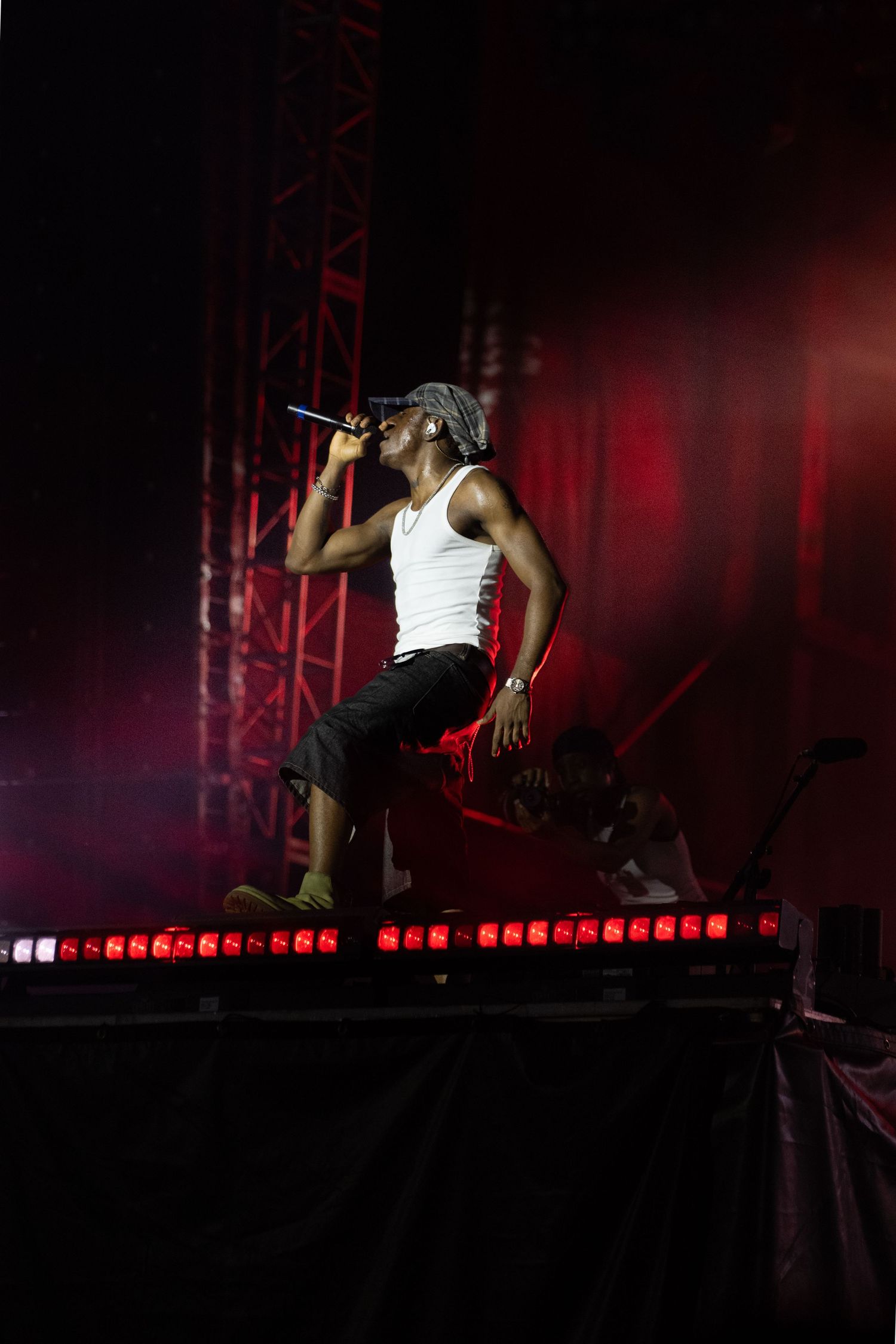 Male performer in white tank top sings into microphone on stage with red lighting at outdoor concert.