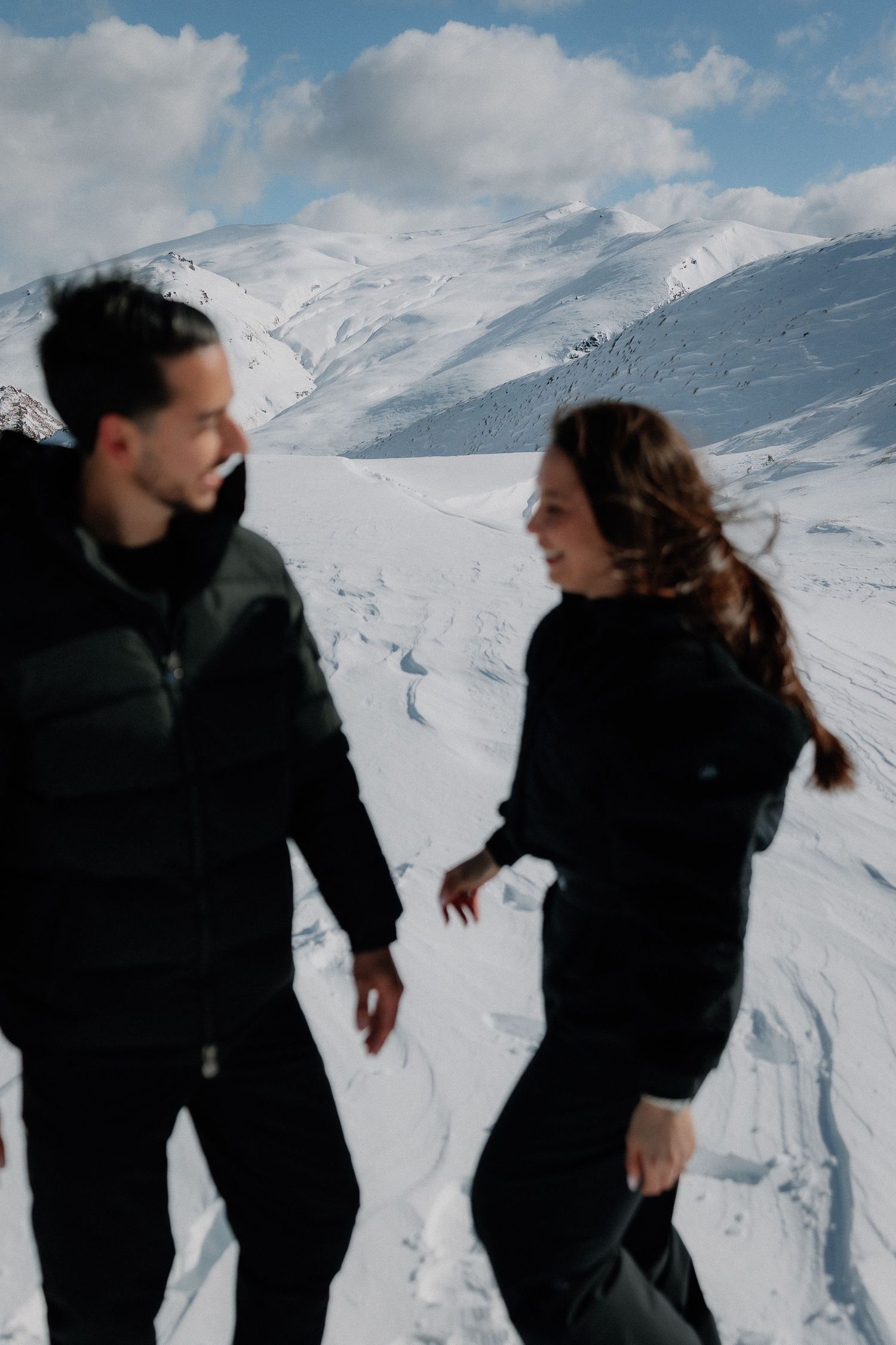 Couple holding hands while gazing at snow-covered mountain peaks under a cloudy sky in winter.