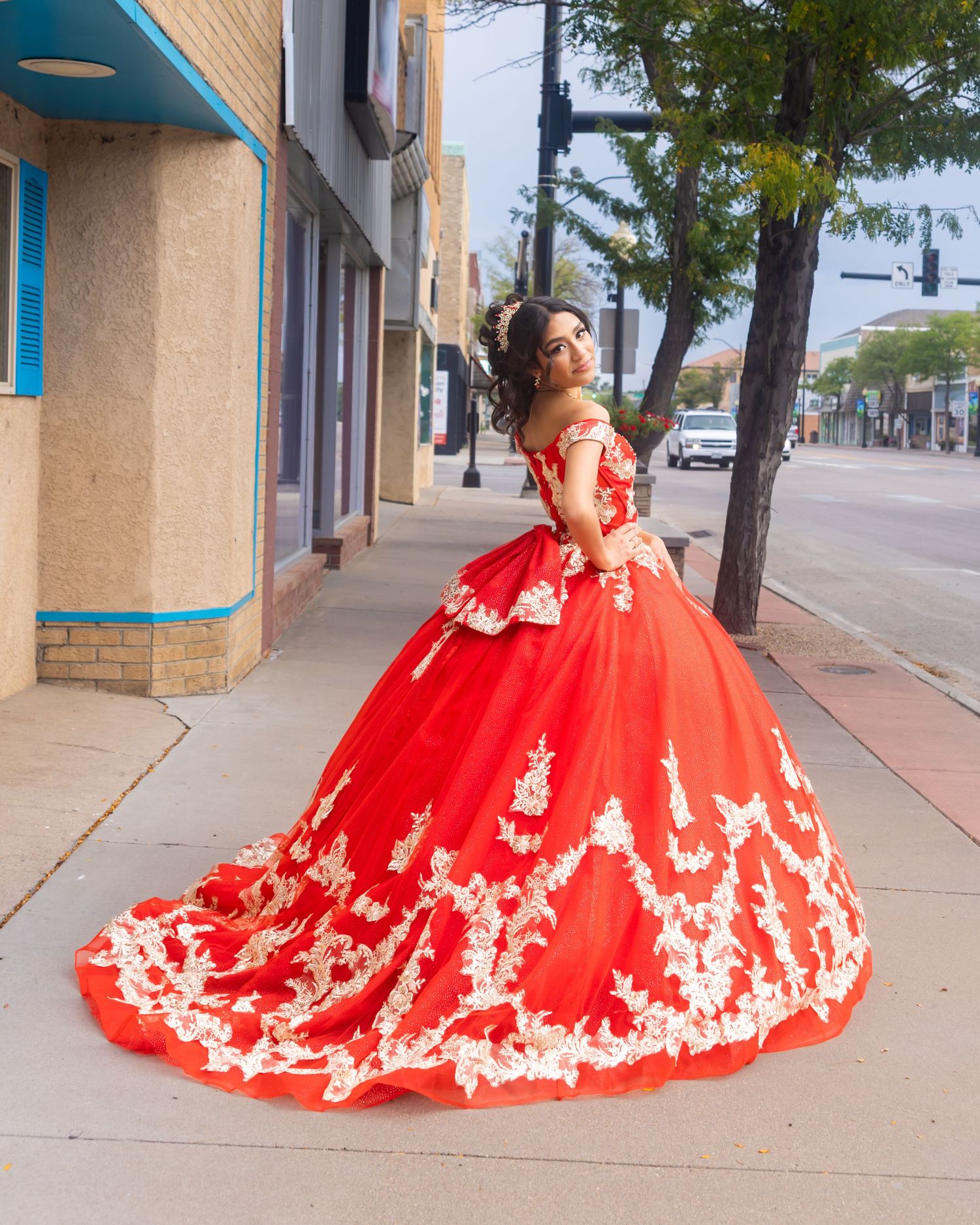 Young woman posing outdoors in a stunning red ball gown with white lace appliques on a city sidewalk.