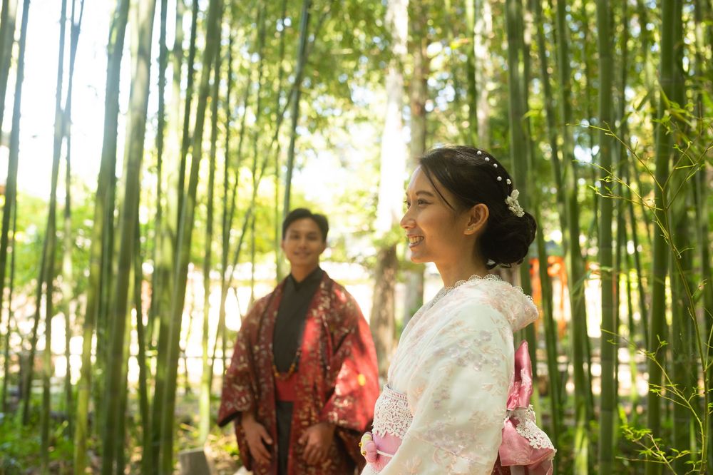 Two women in traditional Japanese kimono smile and stand among tall green bamboo stalks in a sunlit forest grove.