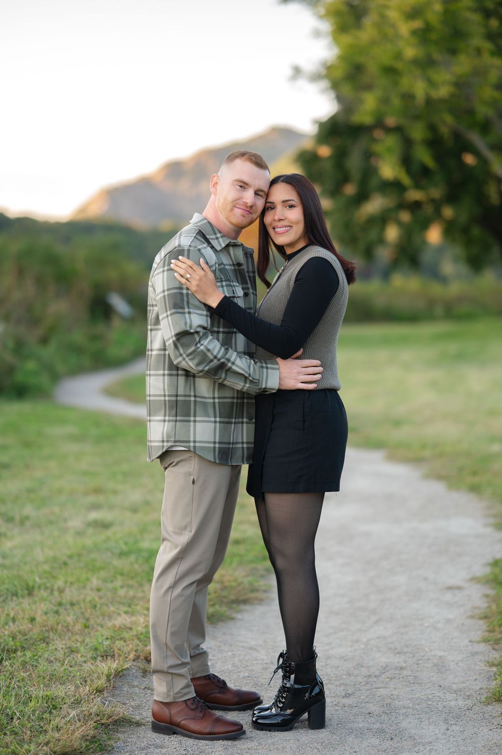 A smiling engaged couple embraces on a gravel path surrounded by green grass with mountains glowing in soft evening light.