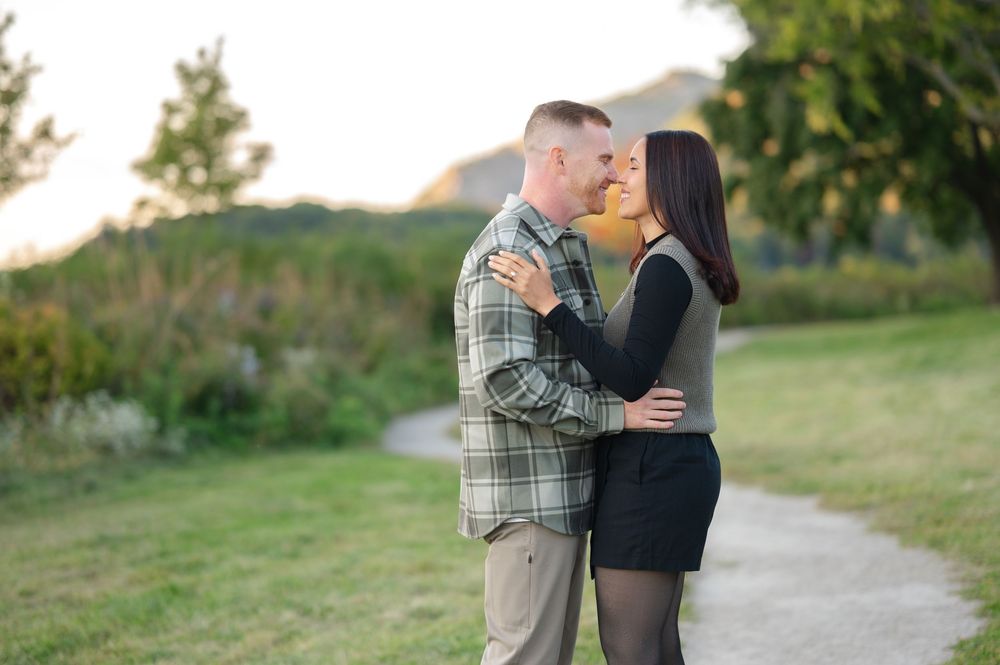 Couple embracing and smiling on a winding gravel path in a grassy park with trees and hills in the background at sunset.