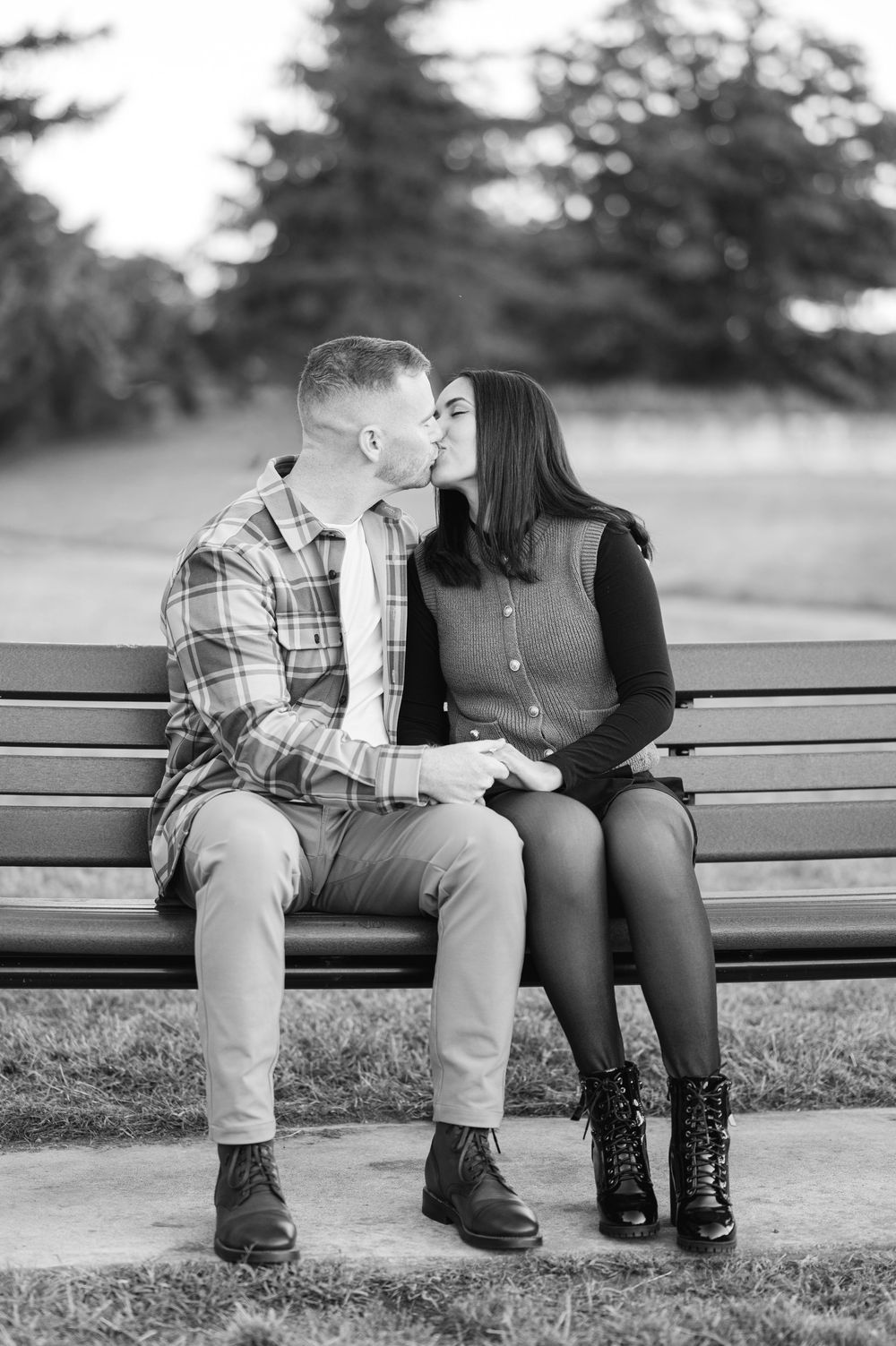 Black and white photo of a couple kissing on a park bench, holding hands, with trees in the background.