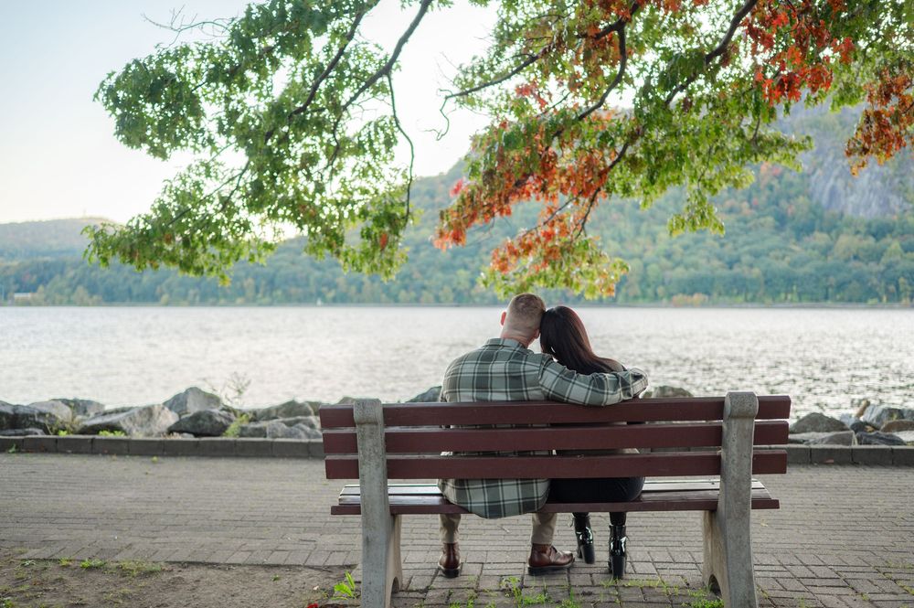 Couple sitting close together on a wooden bench under autumn foliage, overlooking a calm river with forested hills in the distance.