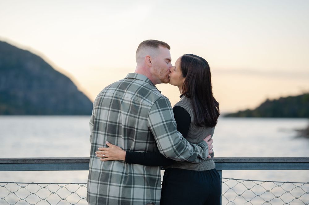 Couple in an embrace sharing a kiss on a waterfront railing at sunset with calm water and hazy mountains in the background.