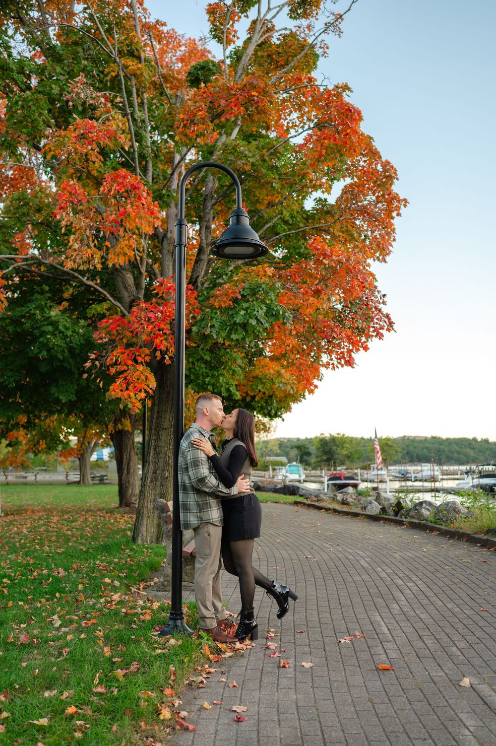 A couple kisses beside a black lamp post on a brick lakeside path under a tree with bright orange autumn foliage.