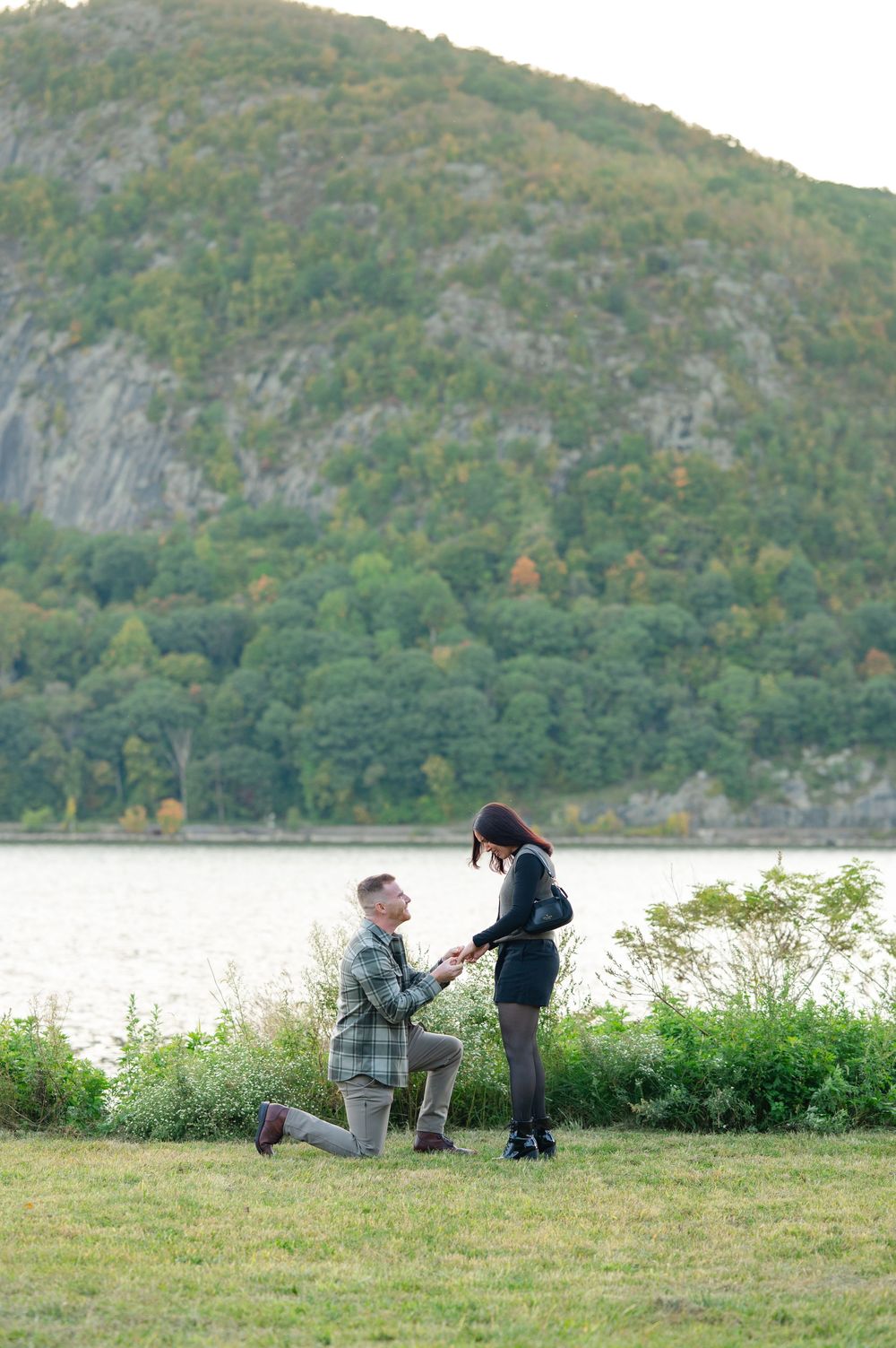 A man kneels on one knee proposing to his girlfriend on a grassy lawn by a river with a forested mountain behind them.