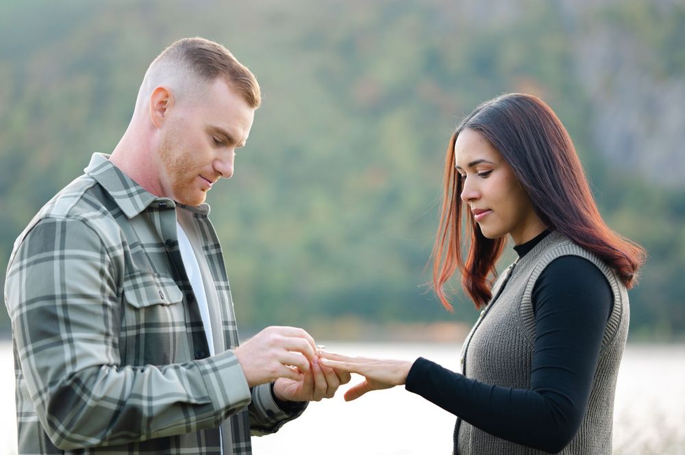 A man in a plaid flannel shirt places a ring on a woman's finger during an outdoor proposal with a scenic lake and mountains behind them.