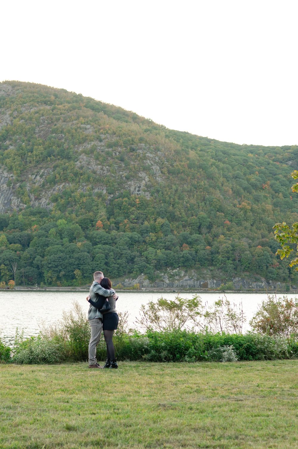 Couple embracing on a grassy riverbank with a forested mountain rising across the water in autumn.