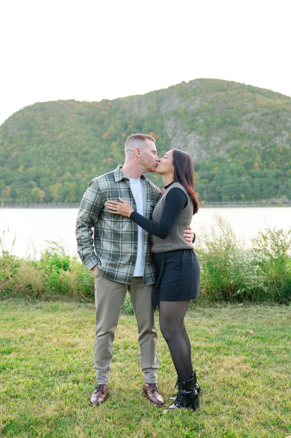 Couple kissing on a grassy lawn by a calm river with a forested mountain behind them, he in a plaid shirt and she in a skirt and boots.