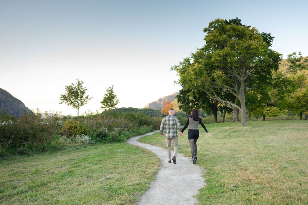 Couple walking along a winding dirt path through a grassy meadow with trees and distant hills at sunset.