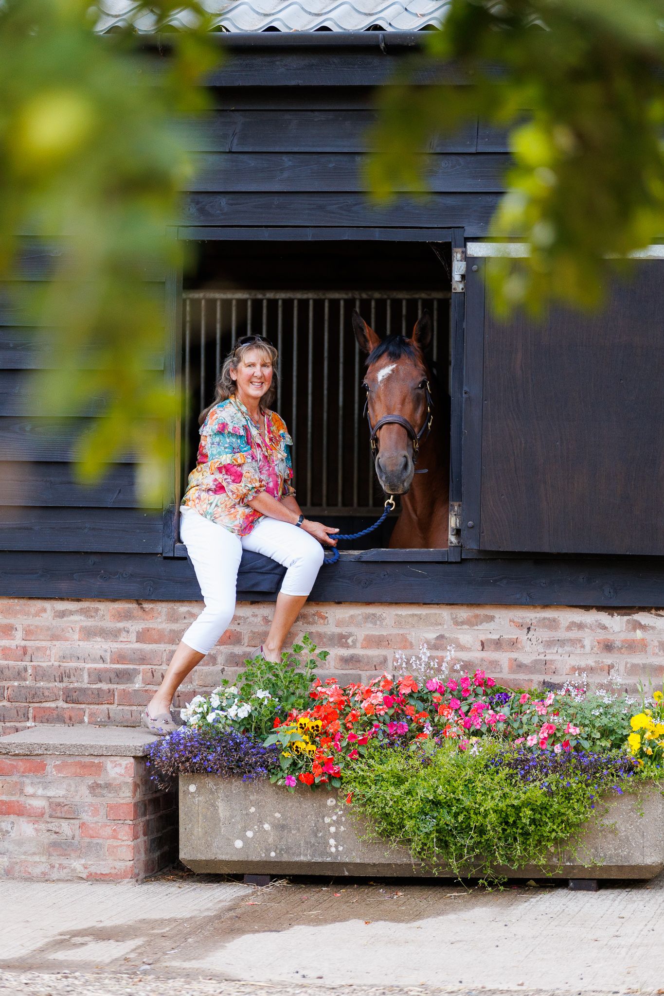 Woman in a long navy blue dress walking a bay horse along a quiet country lane lined with green trees and golden grass.